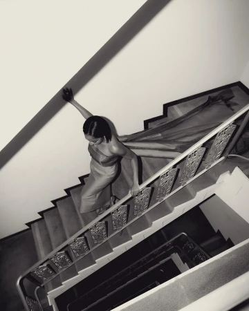 Woman in dress climbs an indoor staircase with ornate metal railing. Black and white photography with high-angle perspective.