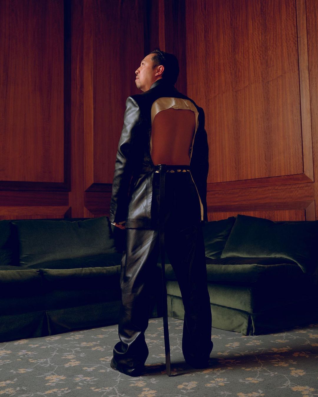 An adult male stands in a black outfit and faces away, indoors with wood-panelled walls and a velvet sofa in the background.