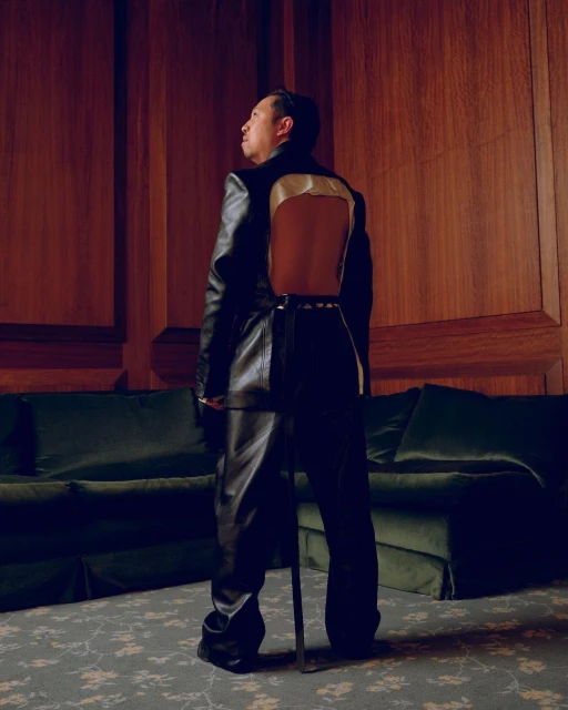 An adult male stands in a black outfit and faces away, indoors with wood-panelled walls and a velvet sofa in the background.
