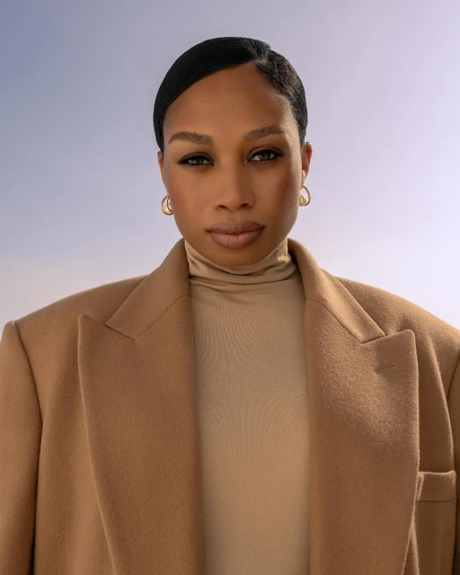 Headshot of a Black woman wearing a tan blazer and turtleneck against a soft blue background, looking directly at the camera.