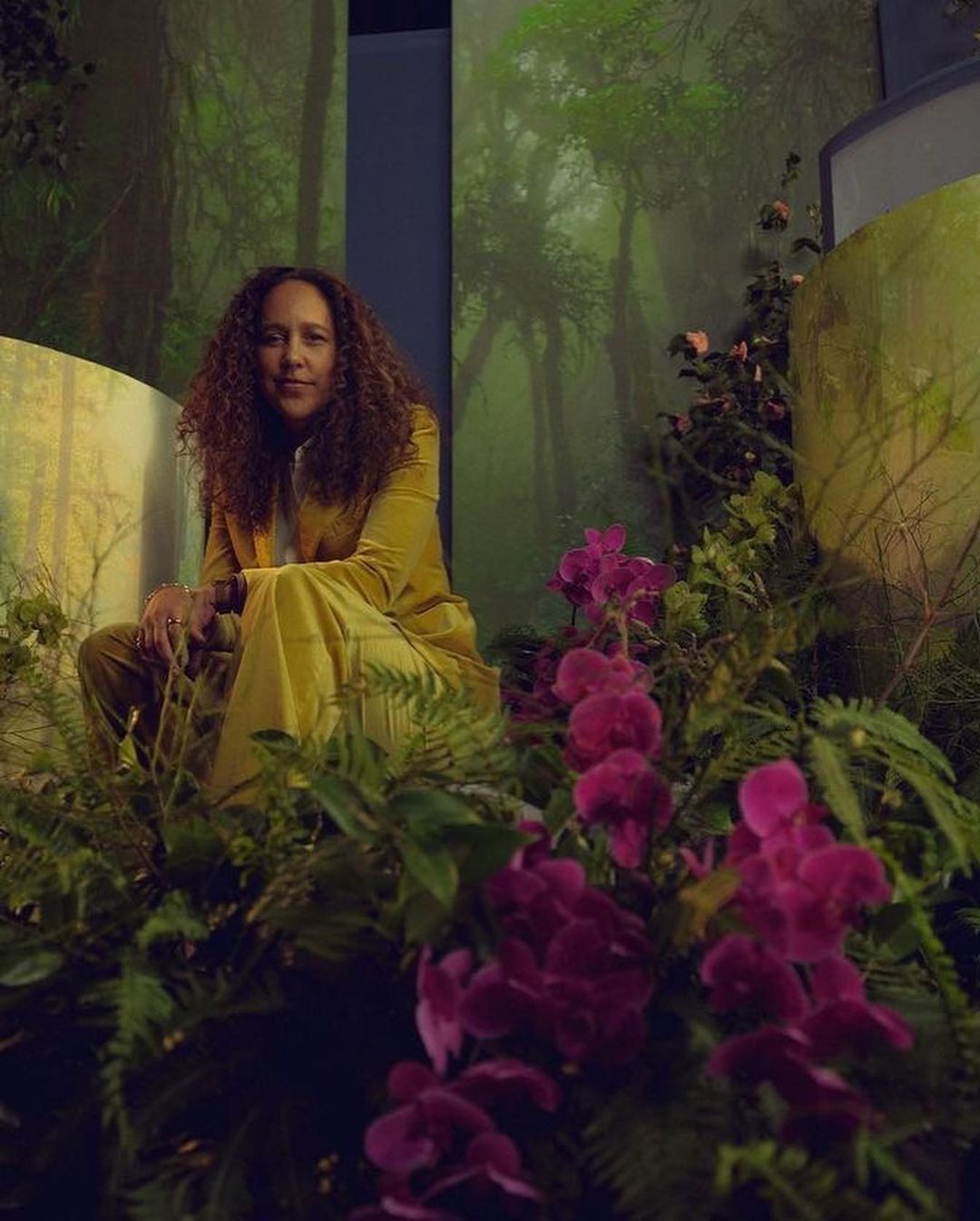 Woman with curly hair wearing a mustard-colored suit, seated amongst lush greenery and bright magenta orchids with a forest backdrop.