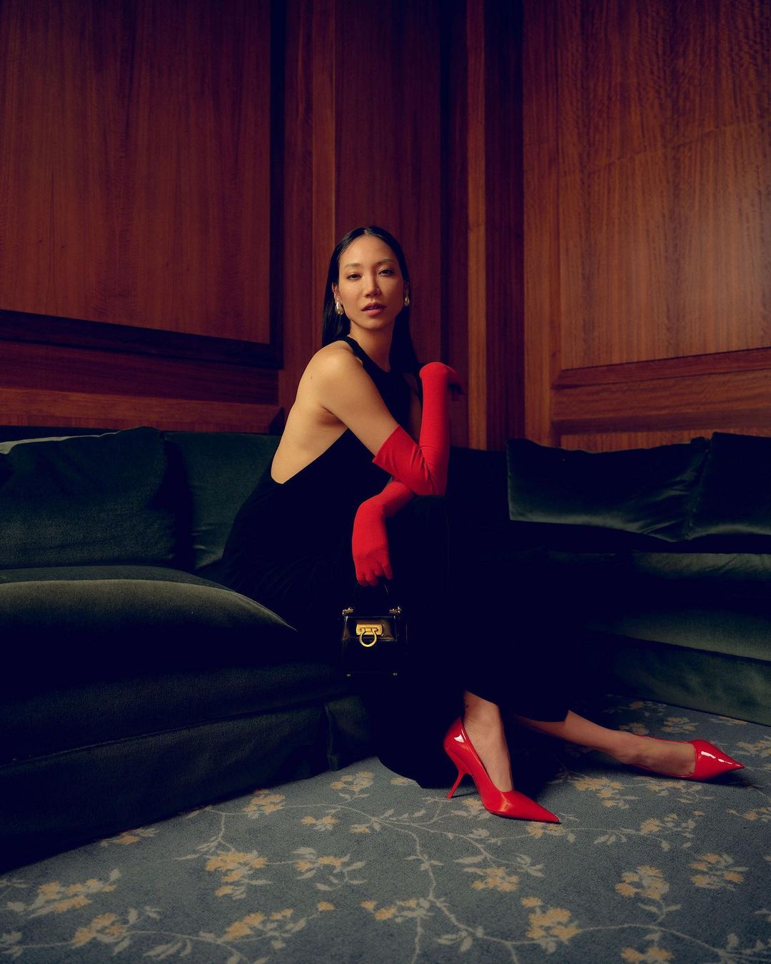 Elegant Asian woman poses indoors on a green sofa in a black dress, red gloves and heels, holding a structured black handbag.
