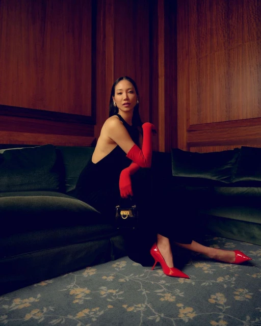Elegant Asian woman poses indoors on a green sofa in a black dress, red gloves and heels, holding a structured black handbag.