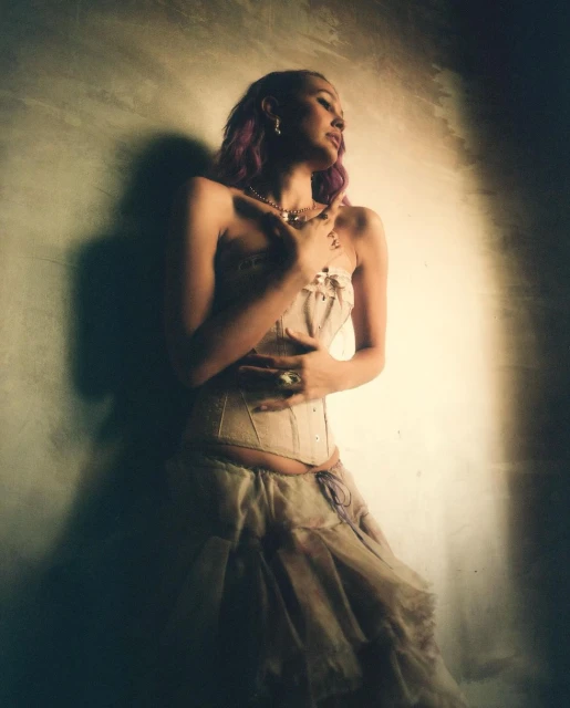 Woman in corset poses near textured wall in photo studio. She has purple hair and wears jewelry. The image is well-lit and in portrait.