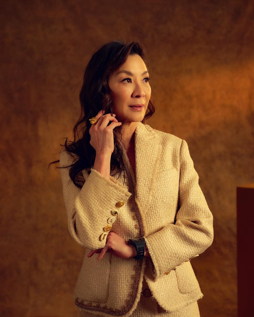 Michelle Yeoh in a portrait. The Malaysian actress wears a tan suit, looking off to the left against a mottled brown studio background.