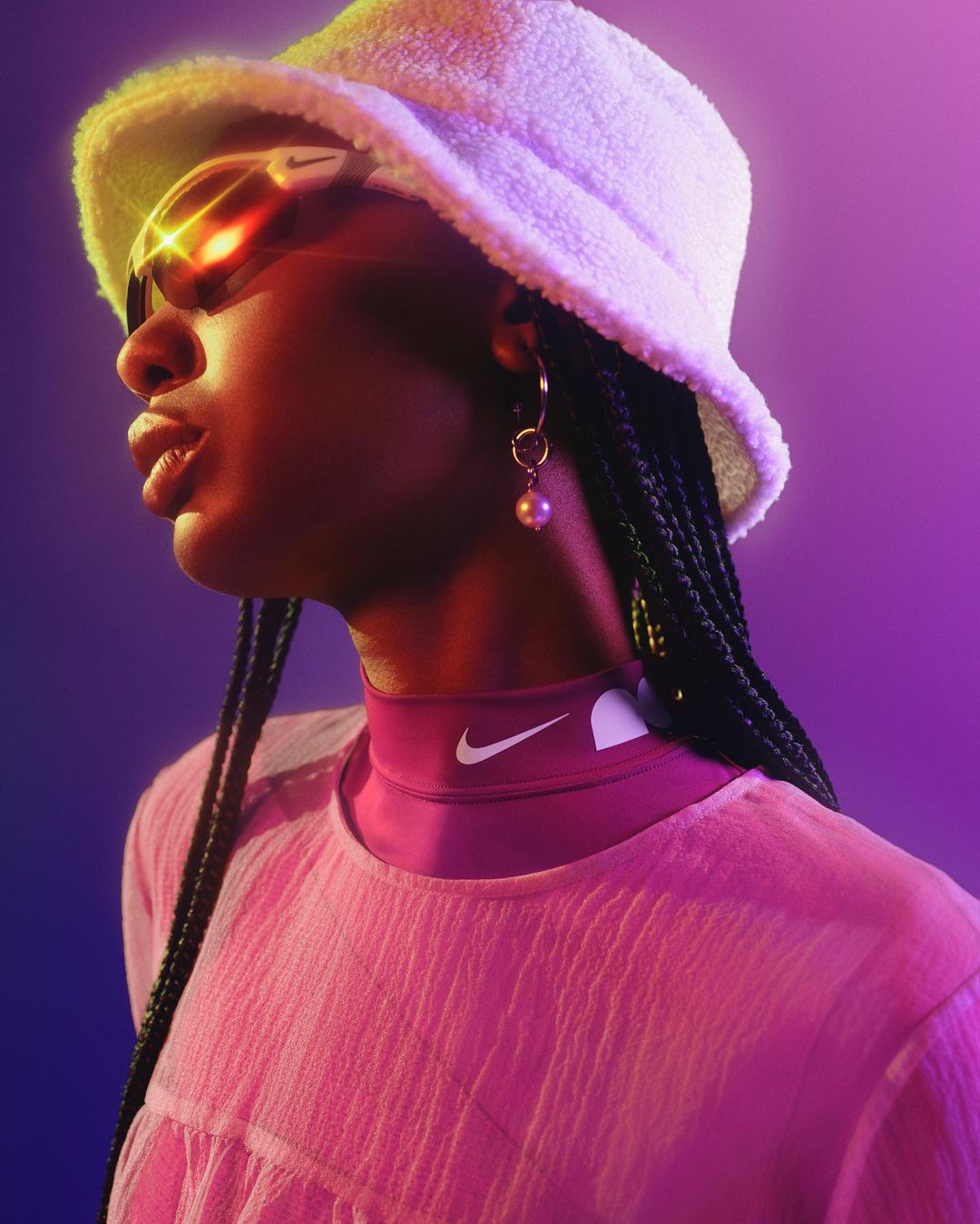 Woman with braided hair poses wearing a white bucket hat, sunglasses, and a pink collared shirt against a vibrant purple background.