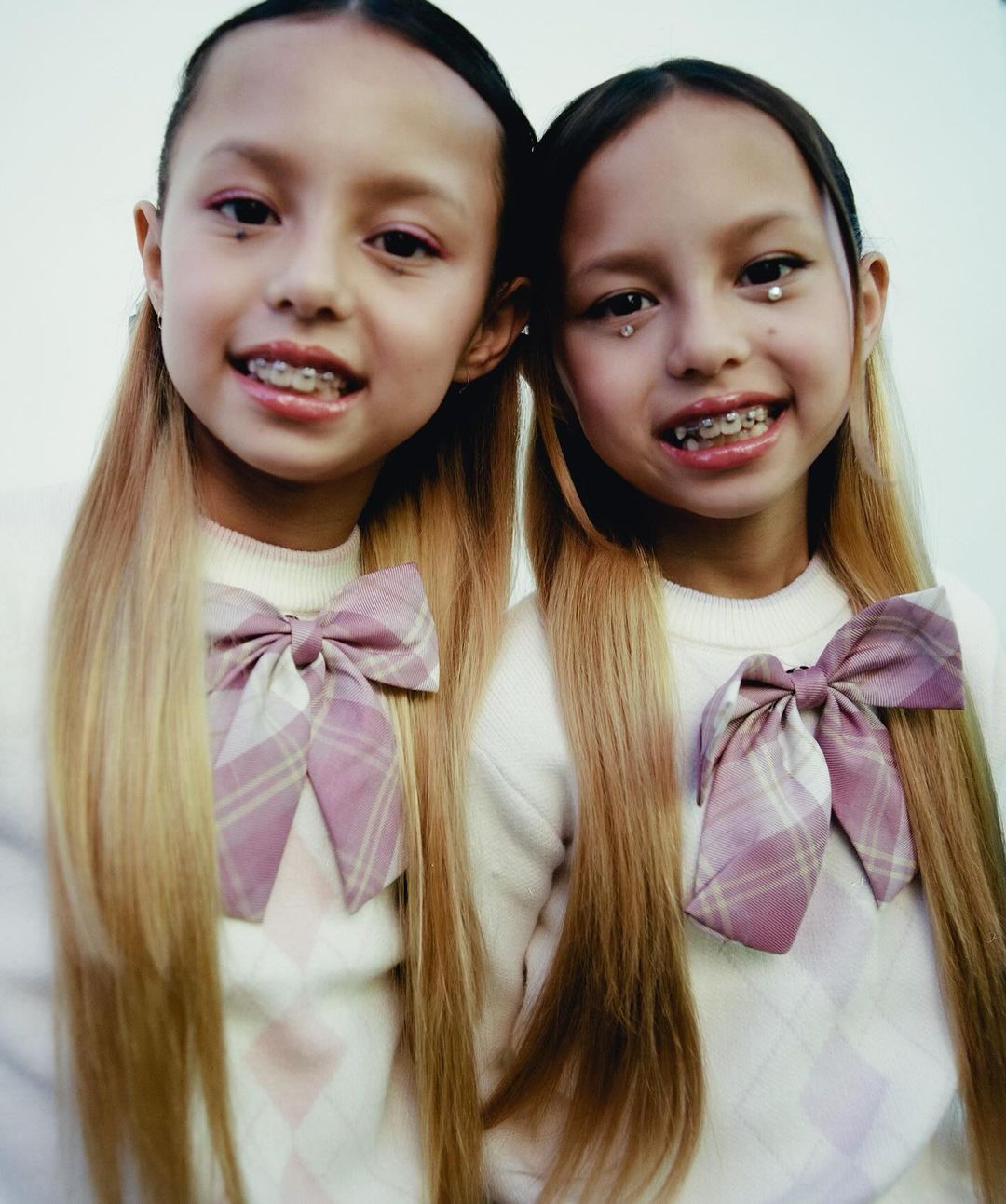 Portrait of two young girls with matching blonde hair, argyle patterned sweaters, and bows smiling into the camera on white background.