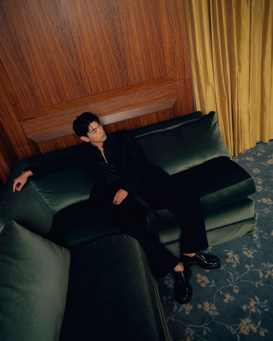 Young man in a black suit poses reclining on a green velvet couch against a wood wall with floral carpet in a warm-lit, indoor setting.