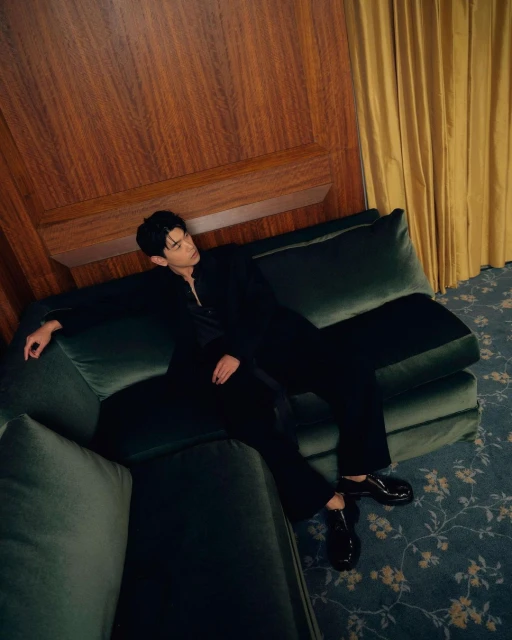 Young man in a black suit poses reclining on a green velvet couch against a wood wall with floral carpet in a warm-lit, indoor setting.