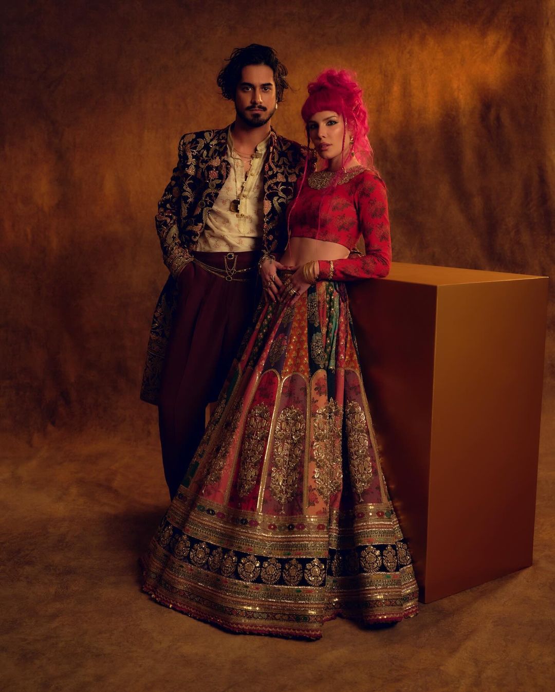 A stylish couple poses indoors against a neutral backdrop; the woman wears a colorful, patterned lehenga and the man a patterned jacket.