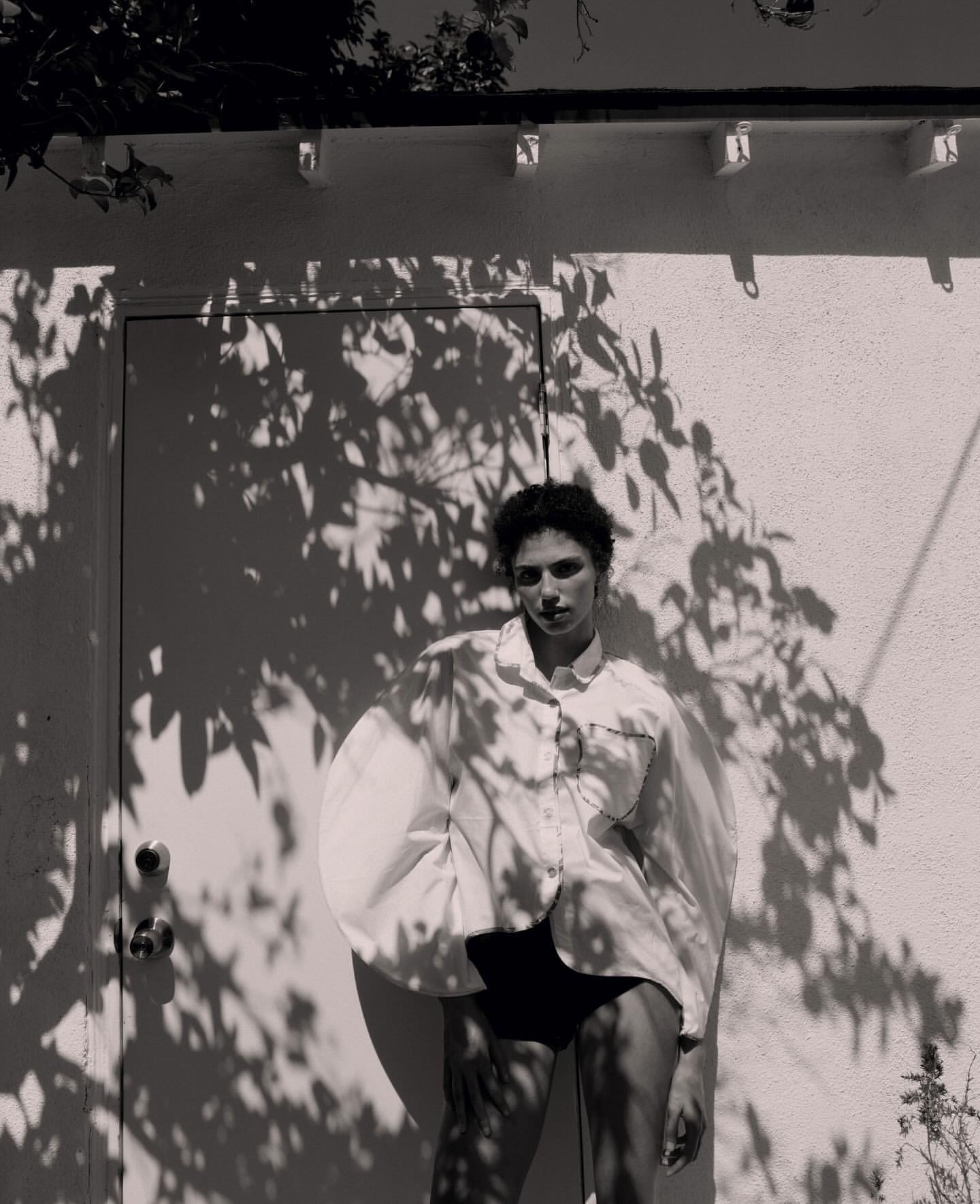 Monochrome fashion shot of a woman in an oversized white shirt and black bottoms, posing in front of a white building with shadows.