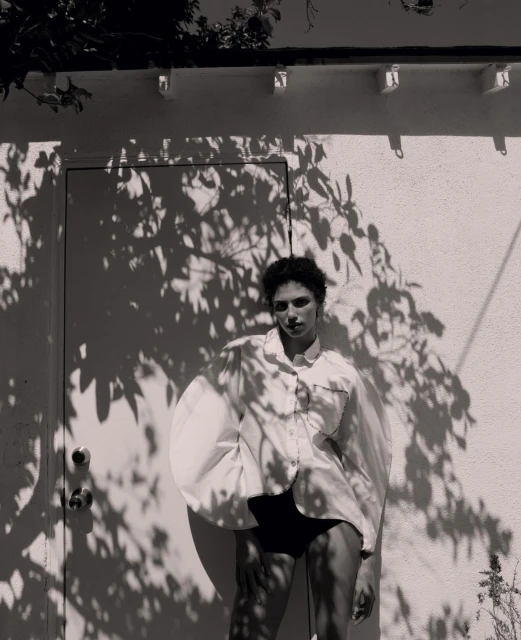 Monochrome fashion shot of a woman in an oversized white shirt and black bottoms, posing in front of a white building with shadows.
