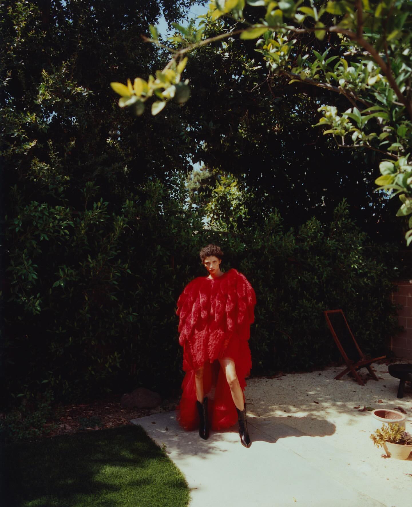 A fashion shot in a garden setting features a woman in a red, ruffled coat and black boots on a paved area surrounded by greenery.