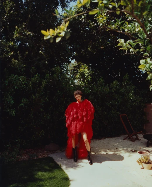 A fashion shot in a garden setting features a woman in a red, ruffled coat and black boots on a paved area surrounded by greenery.