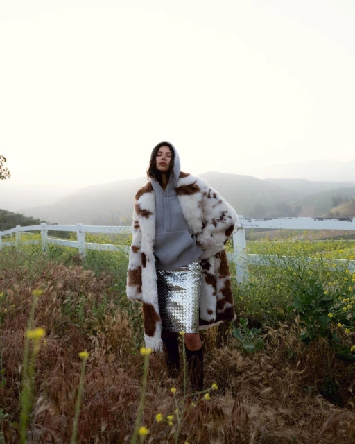 A woman in a fur coat and silver skirt stands in a grassy field with a white fence and distant hills under a bright, hazy sky.