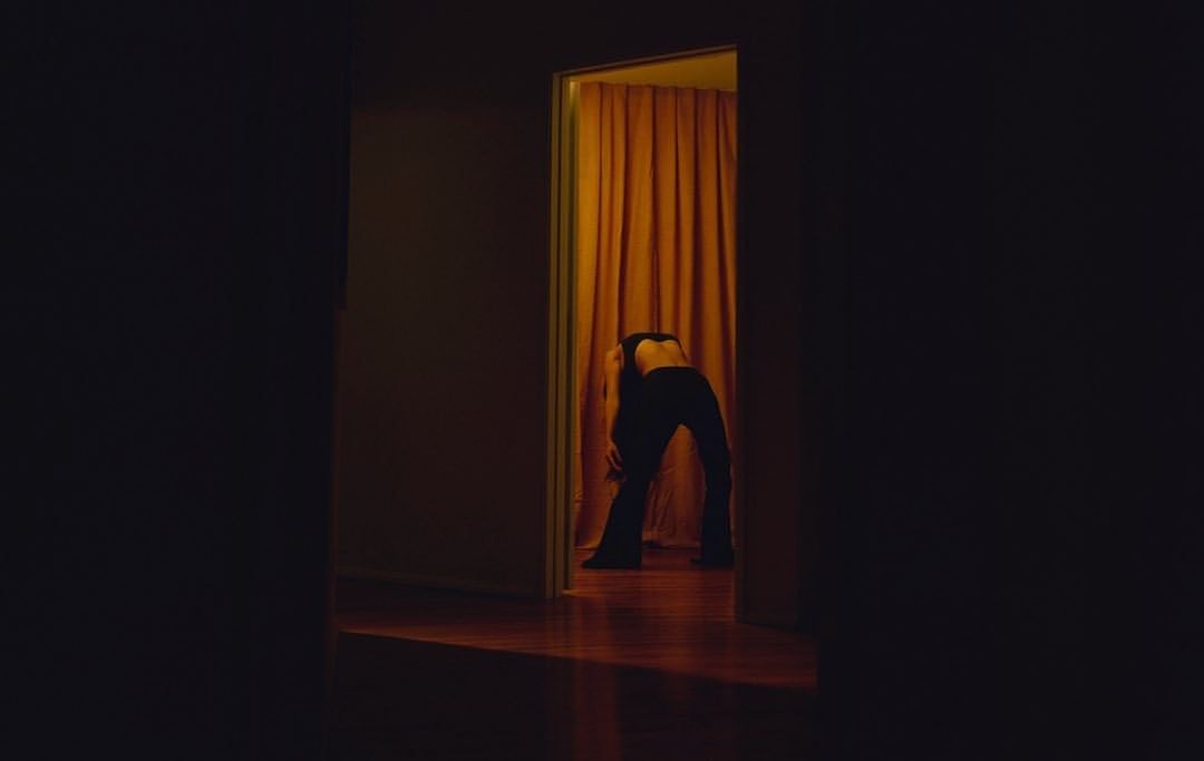 Woman bends backward inside a doorway with orange curtain, silhouetted in dark clothing against golden light, indoor setting.