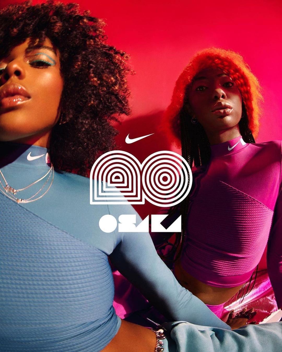 Two women with natural hair pose in blue and magenta long-sleeve tops in a studio with a geometric Nike logo.