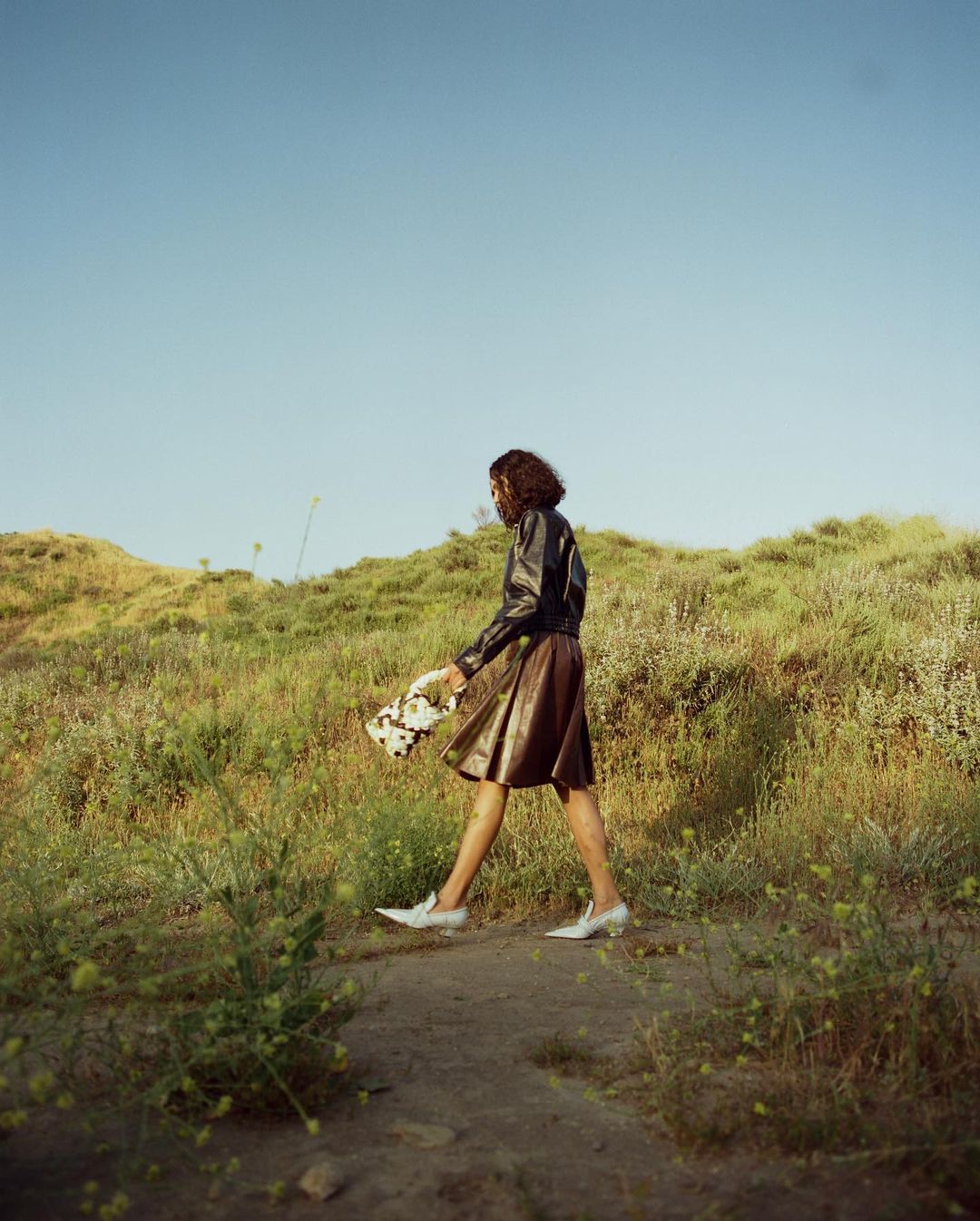 Woman walking on a grassy outdoor path wearing a dark leather jacket, brown skirt, and white shoes, photographed by Kanya Iwana.