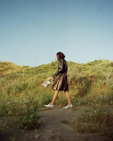 Woman walking on a grassy outdoor path wearing a dark leather jacket, brown skirt, and white shoes, photographed by Kanya Iwana.