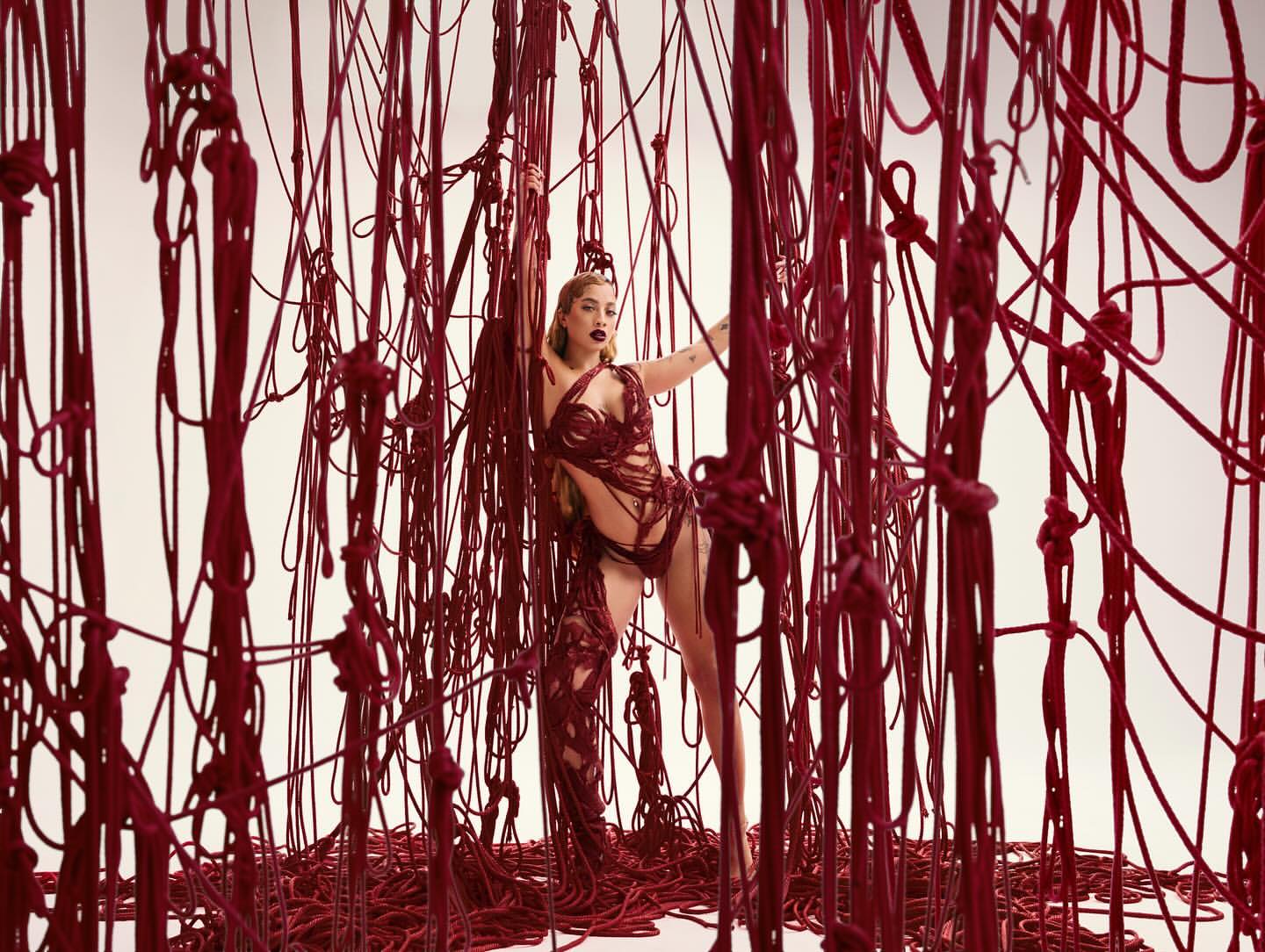 Artistic fashion shot of a woman posing among a curtain of red rope against a white backdrop in a studio; wearing an avant-garde outfit.