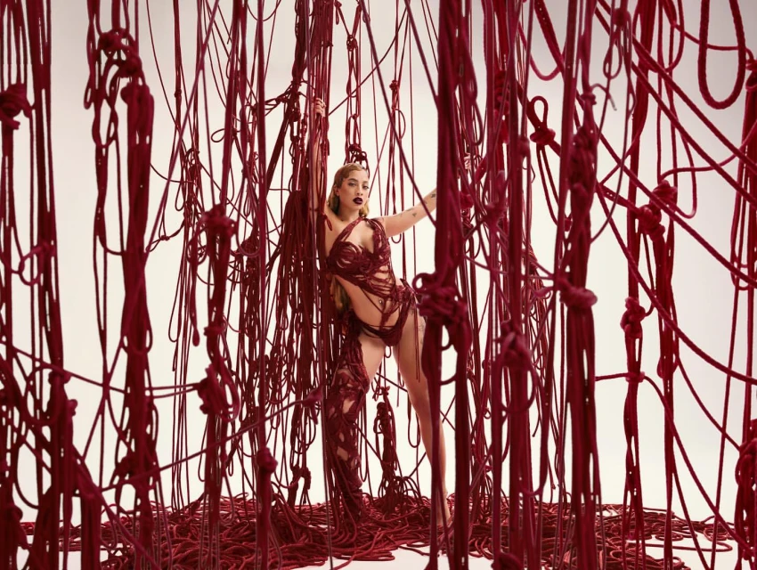 Artistic fashion shot of a woman posing among a curtain of red rope against a white backdrop in a studio; wearing an avant-garde outfit.
