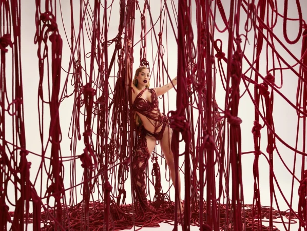 Artistic fashion shot of a woman posing among a curtain of red rope against a white backdrop in a studio; wearing an avant-garde outfit.