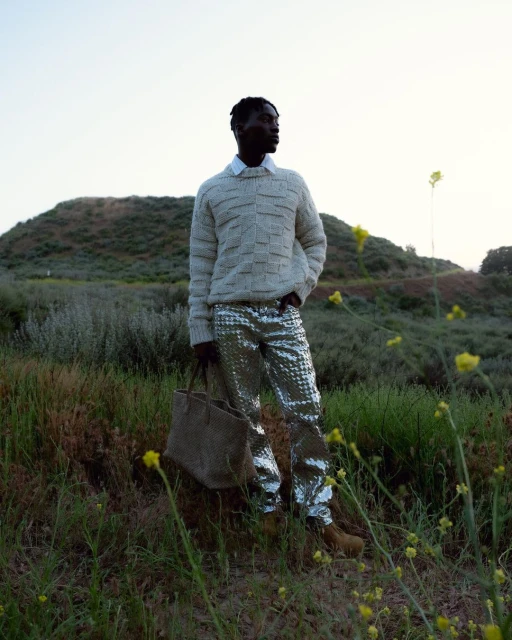 A stylish Black man stands in a grassy field wearing a knit sweater and metallic silver pants, carrying a tote bag on a sunny day.