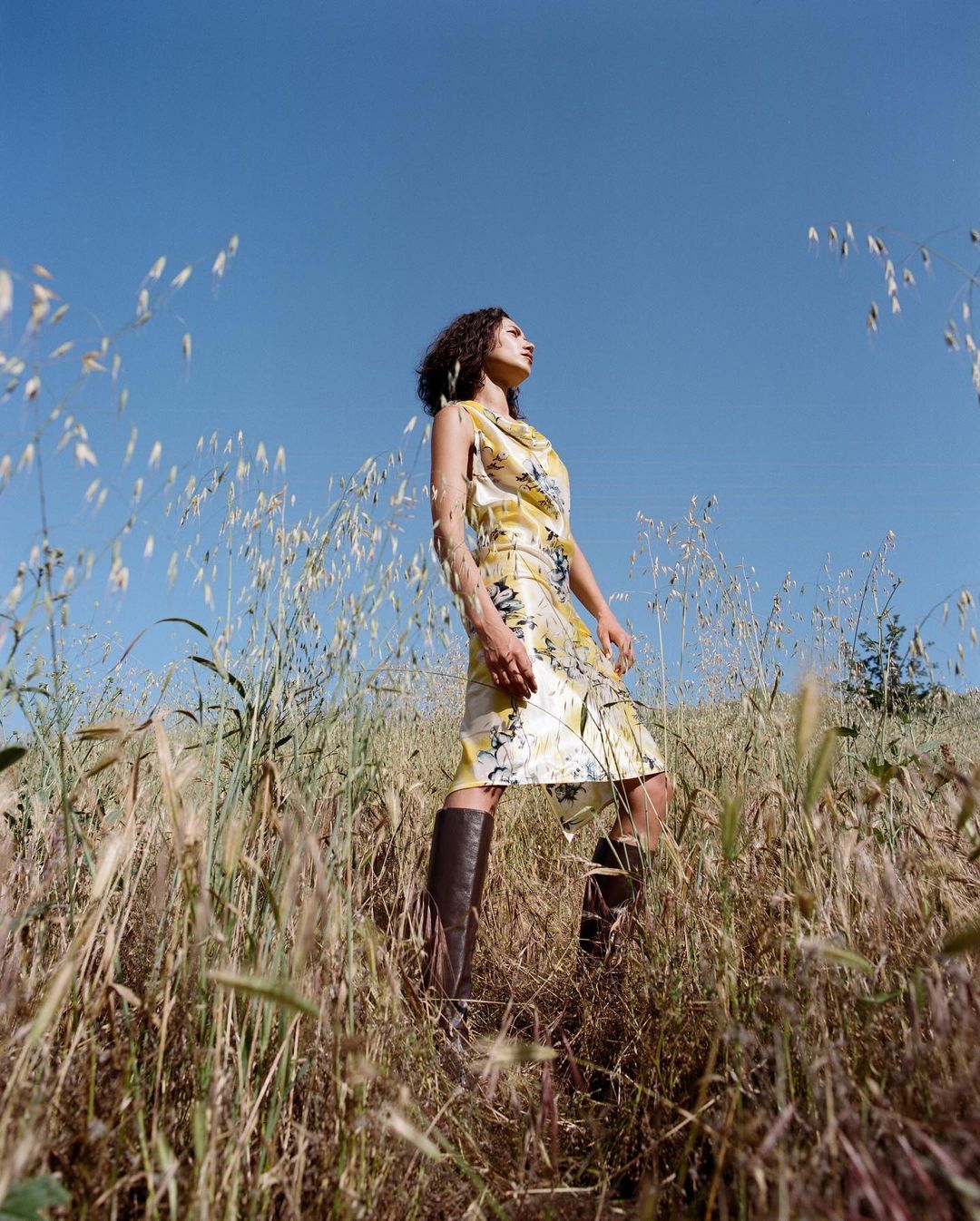 Woman in patterned yellow dress and tall brown boots posing in a field of tall grass under a clear blue sky