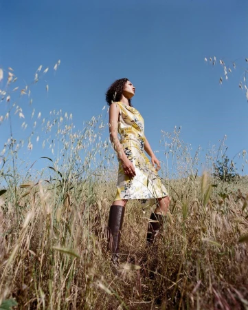 Woman in patterned yellow dress and tall brown boots posing in a field of tall grass under a clear blue sky