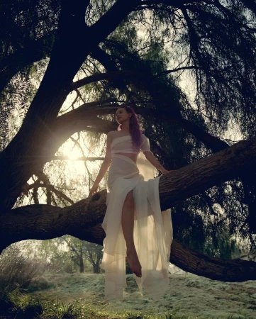 Woman in flowing white dress sits on a large tree branch in a dreamy forest setting with soft light, wearing pastel hair and bare feet.