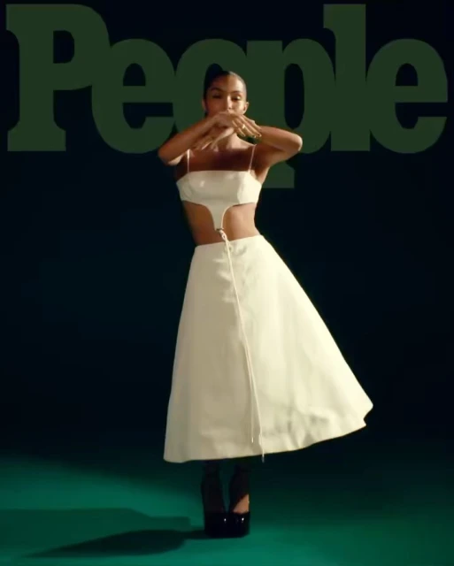 A woman in a white two-piece outfit and black platform heels poses with her hands near her face on a green background for People Magazine.