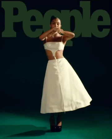 A woman in a white two-piece outfit and black platform heels poses with her hands near her face on a green background for People Magazine.