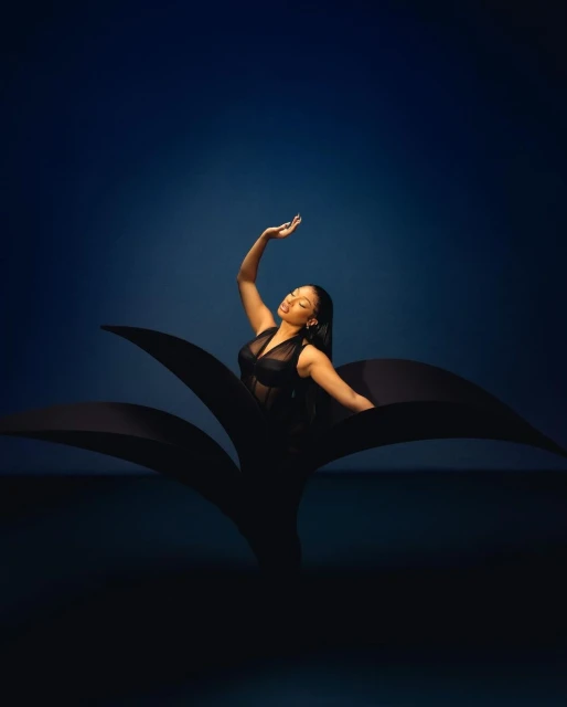 Portrait of a woman in black attire gracefully posing with one arm raised against a dark blue backdrop, framed by stylized black paper.