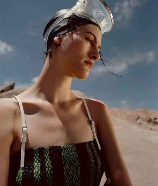 A young woman with dark hair and fair skin poses outdoors in a woven dress and swim goggles in a fashion editorial shot.