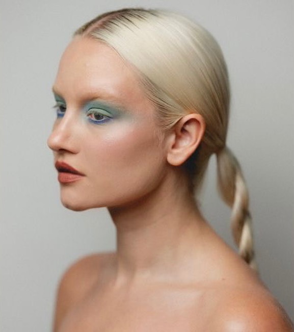 Close-up portrait of a young adult female with platinum blonde hair in a low ponytail, wearing blue and green eye makeup.