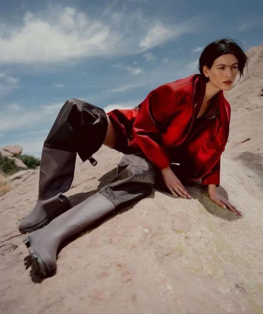 A brunette woman in a red jacket poses on a rock surface in a desert landscape with a cloudy blue sky. She is wearing gray boots.