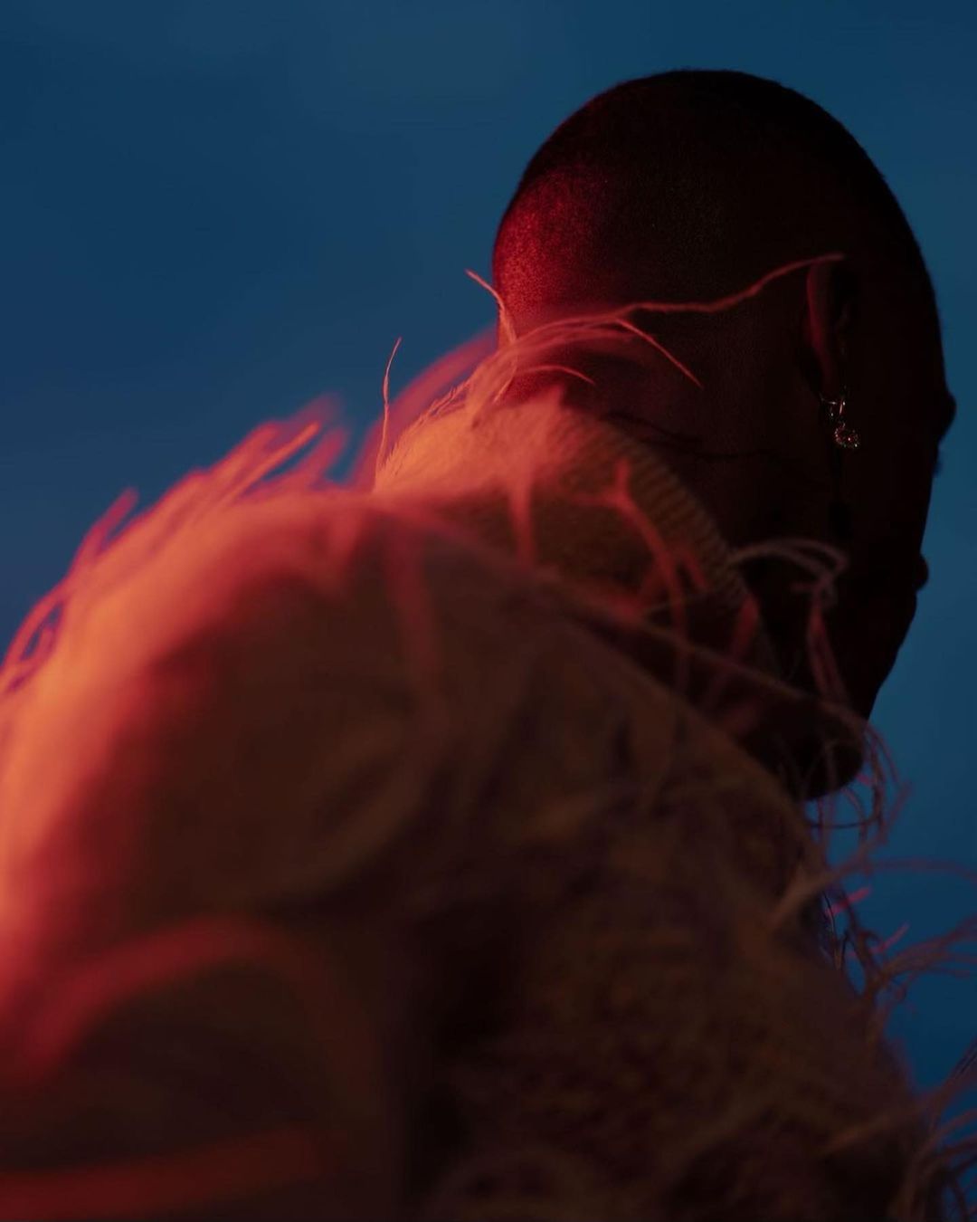 Close-up portrait of a person with a shaved head wearing a feathered garment under blue and red light by Kanya Iwana