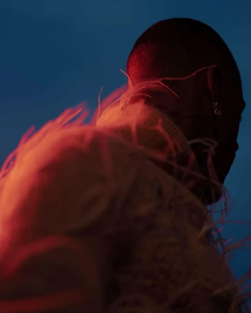 Close-up portrait of a person with a shaved head wearing a feathered garment under blue and red light by Kanya Iwana