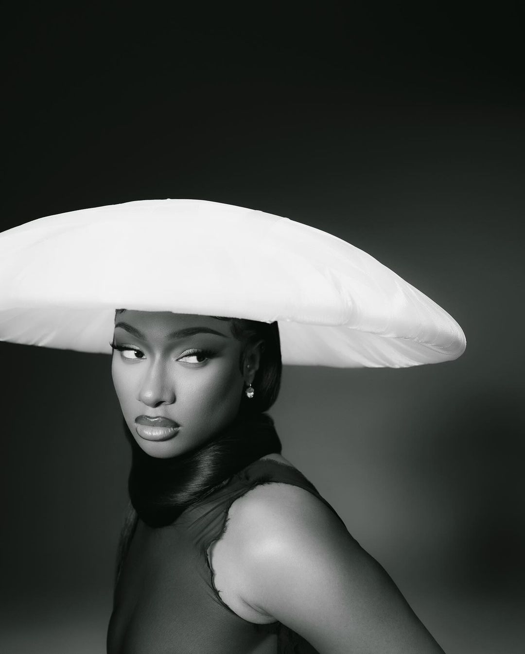 Close-up portrait of a Black woman wearing a large white hat and a sheer burgundy dress, set against a dark gray background.