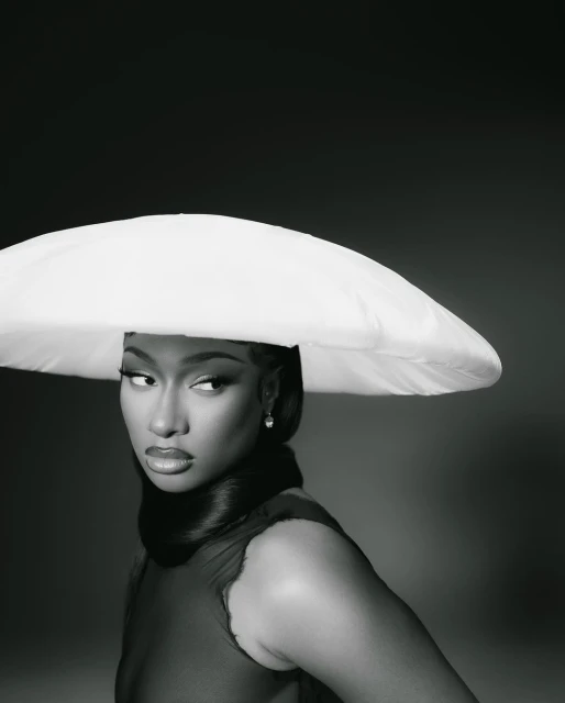 Close-up portrait of a Black woman wearing a large white hat and a sheer burgundy dress, set against a dark gray background.