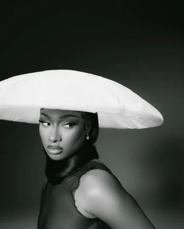 Close-up portrait of a Black woman wearing a large white hat and a sheer burgundy dress, set against a dark gray background.