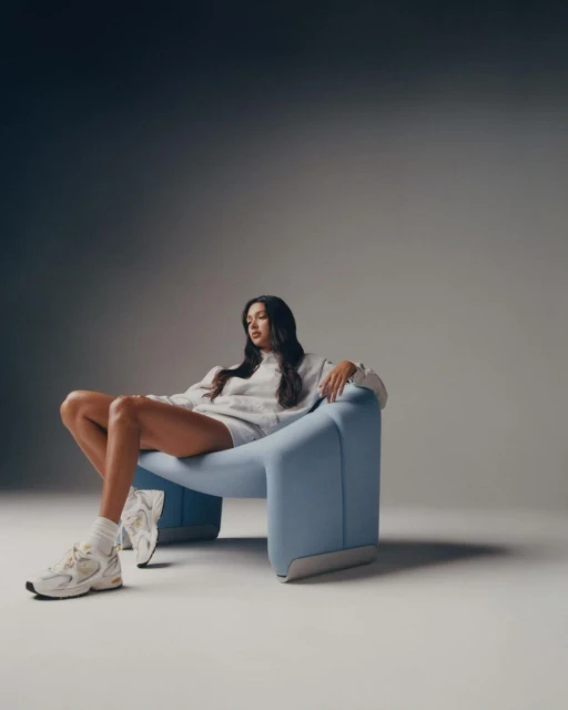 Adult female model wearing a grey sweatshirt and shorts sitting on a light blue chair in a photo studio setting with soft, diffused lighting.