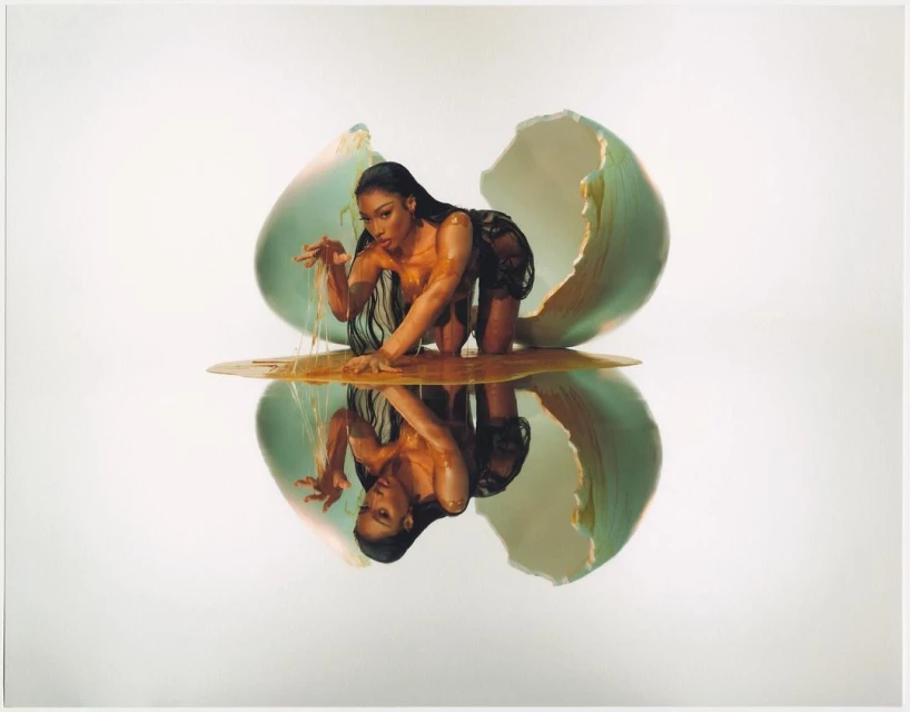 Glamour shot of a woman emerging from a cracked egg, covered in honey, with a reflective surface, and against a white backdrop.