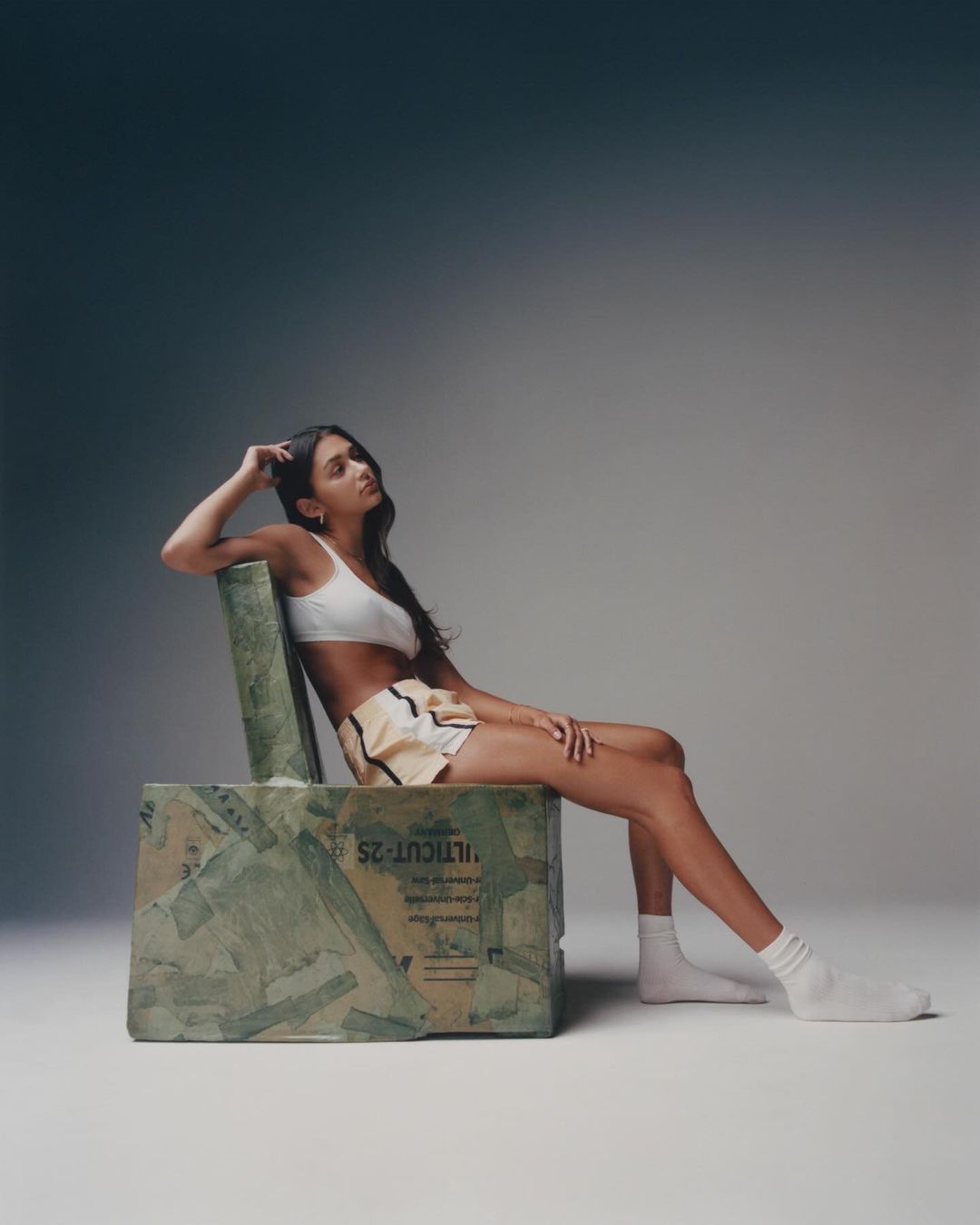 A brunette woman in a white top and shorts poses while sitting on a repurposed cardboard chair against a grey gradient backdrop.