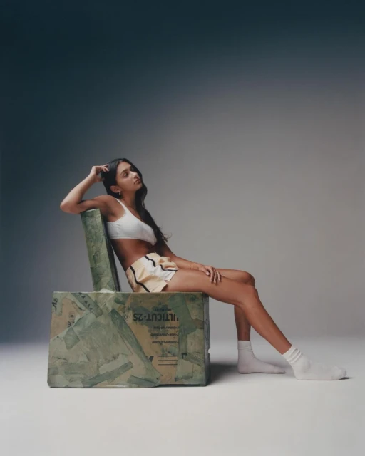 A brunette woman in a white top and shorts poses while sitting on a repurposed cardboard chair against a grey gradient backdrop.