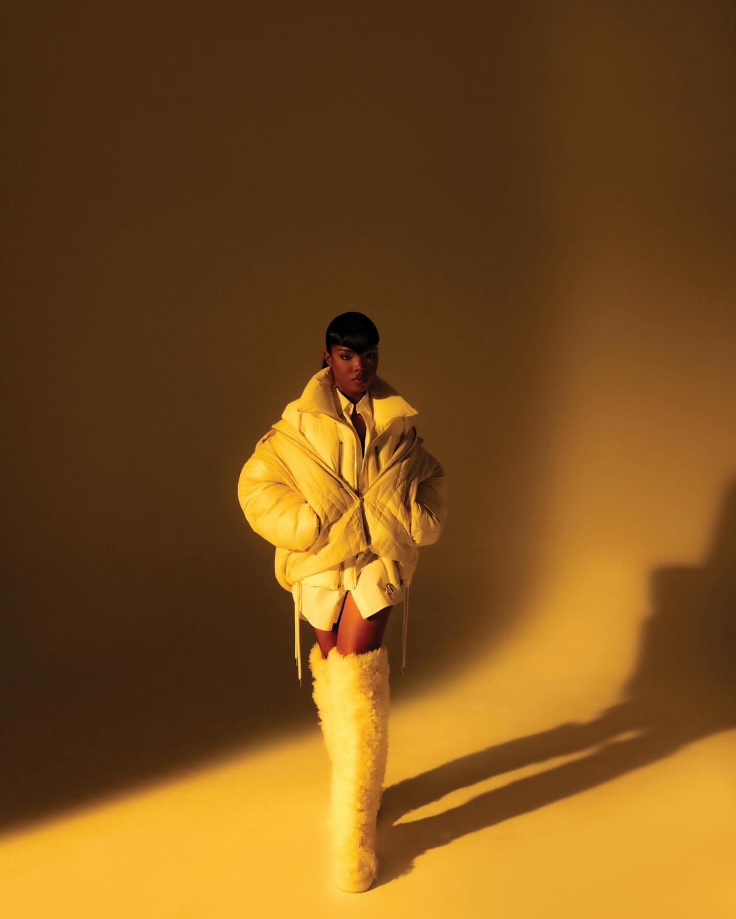 Woman in a yellow puffer jacket poses in a photo studio with a yellow background. She is wearing high, furry yellow boots.