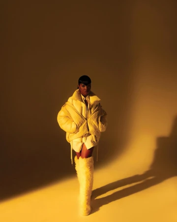 Woman in a yellow puffer jacket poses in a photo studio with a yellow background. She is wearing high, furry yellow boots.