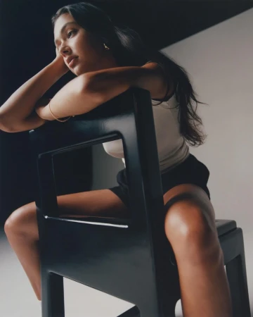 Woman with long dark hair leaning forward over the back of a black wooden chair in a bright studio space.