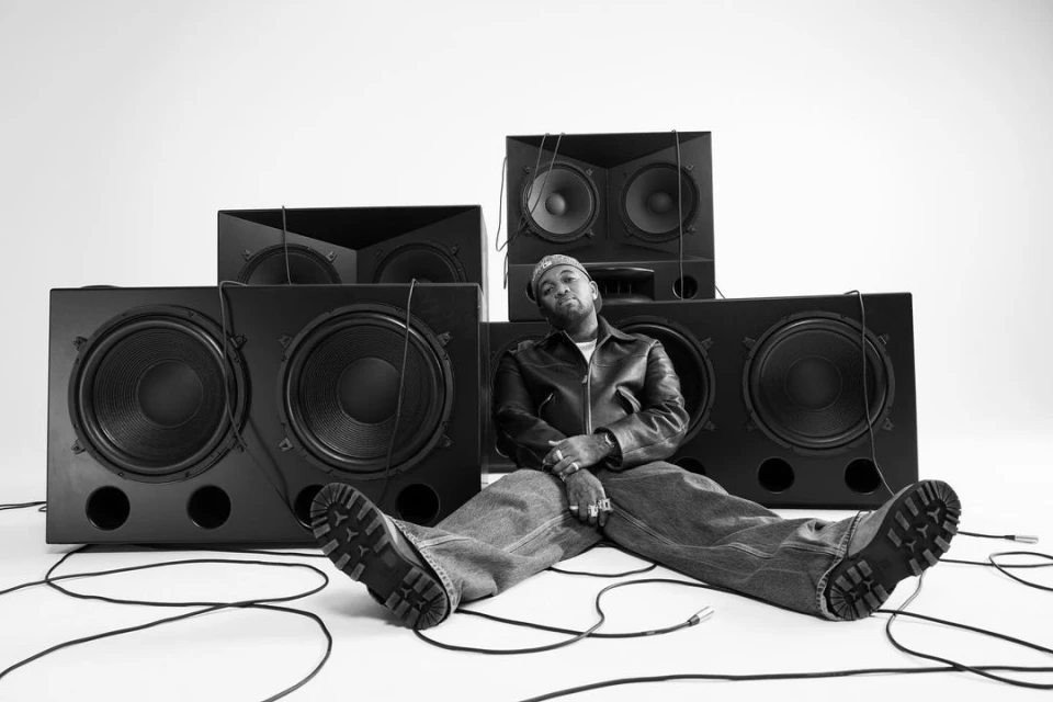 A man wearing a leather jacket and jeans sits with legs outstretched in front of a large stack of black speakers and audio cables against a white background.