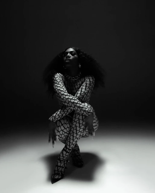 An adult female with dark skin and curly hair is crouched in a studio wearing a printed bodysuit and heels in a dramatic portrait.